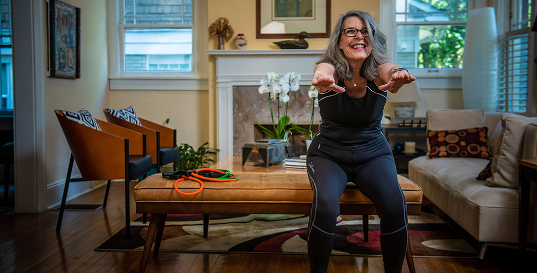 An older woman exercises in her living room with her hands out in front of her while doing a squat.